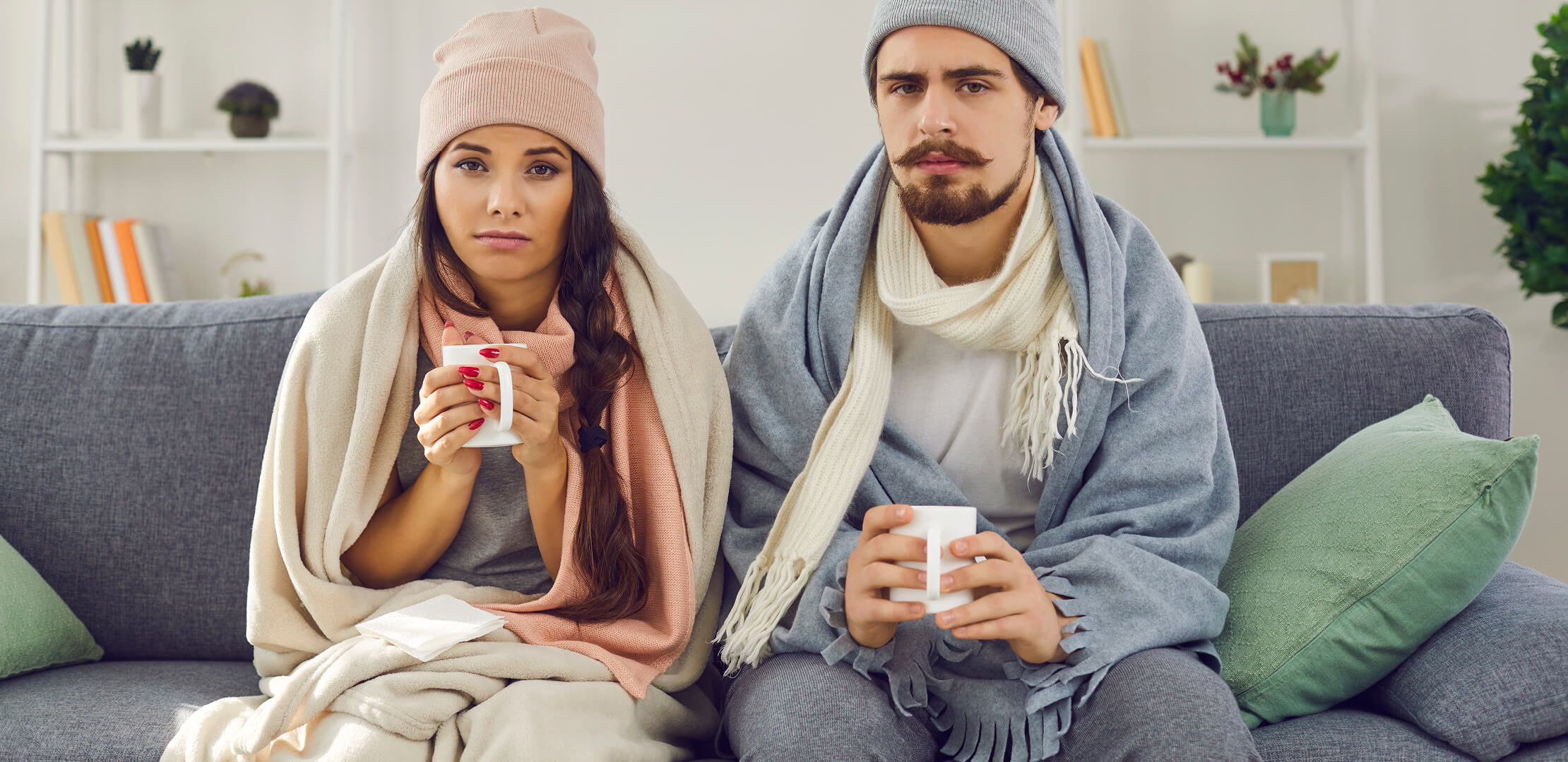Couple bundled up and shivering on couch, holding warm beverages
