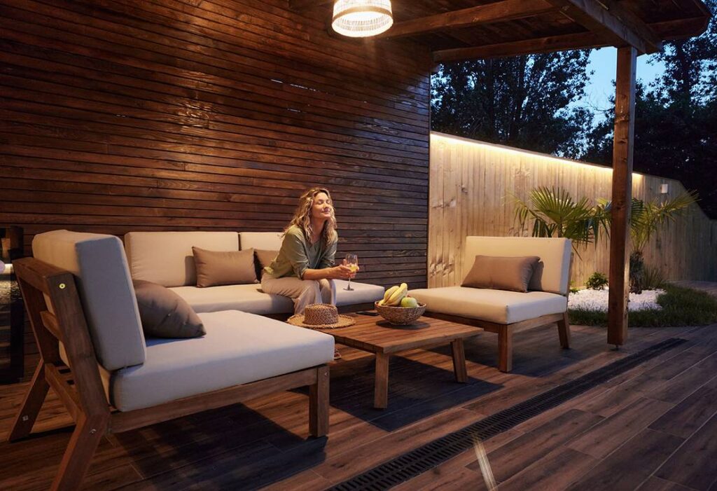 A beautiful young woman is sitting on her dark wooden, modern deck in the evening with a glass of wine in her hands. The decking goes all the way up the wall behind her and there is a pergola covering the deck. She is sitting on modern teak furniture with plush curtains and pillow and there are tropical plants in the background with modern lighting along the fenceline. On the table in front of her is her sun hat and a wicker bowl with bananas and apples.