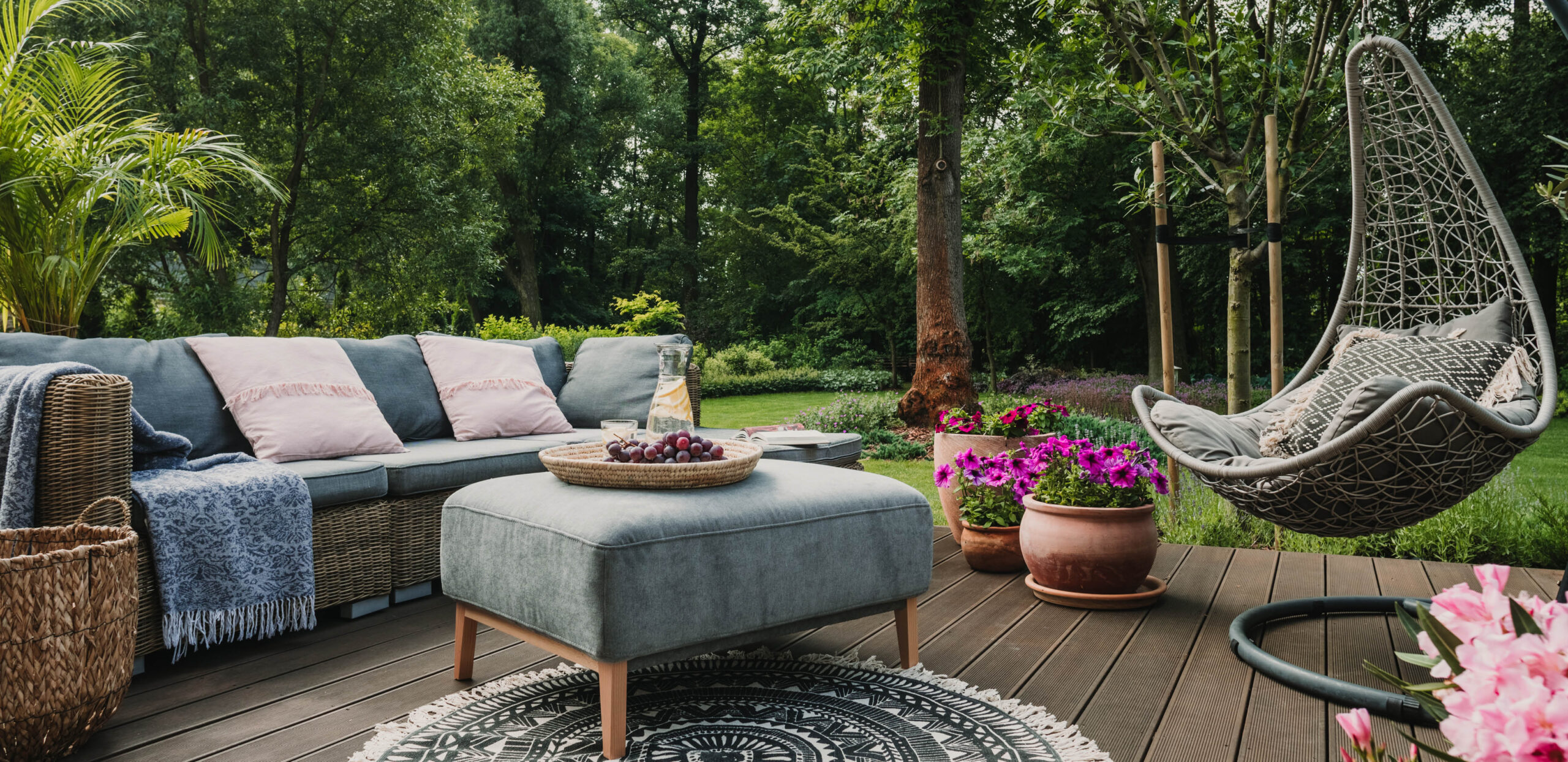 A beautiful, tree-filled back yard with a deck filled with comfortable furniture, a rug and flowers.