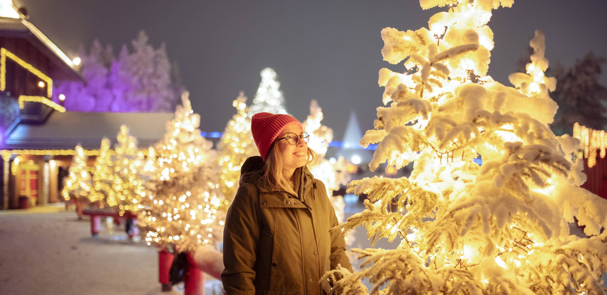 A young woman bundled up in winter clothes, smiling and admiring white holiday lights on evergreen trees leading up to a festive house