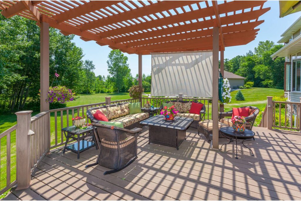 A beautiful deck with lovely furniture and brightly colored pillows and cushions is shaded by a wooden pergola. A lush green lawn, trees and bushes are in the background.