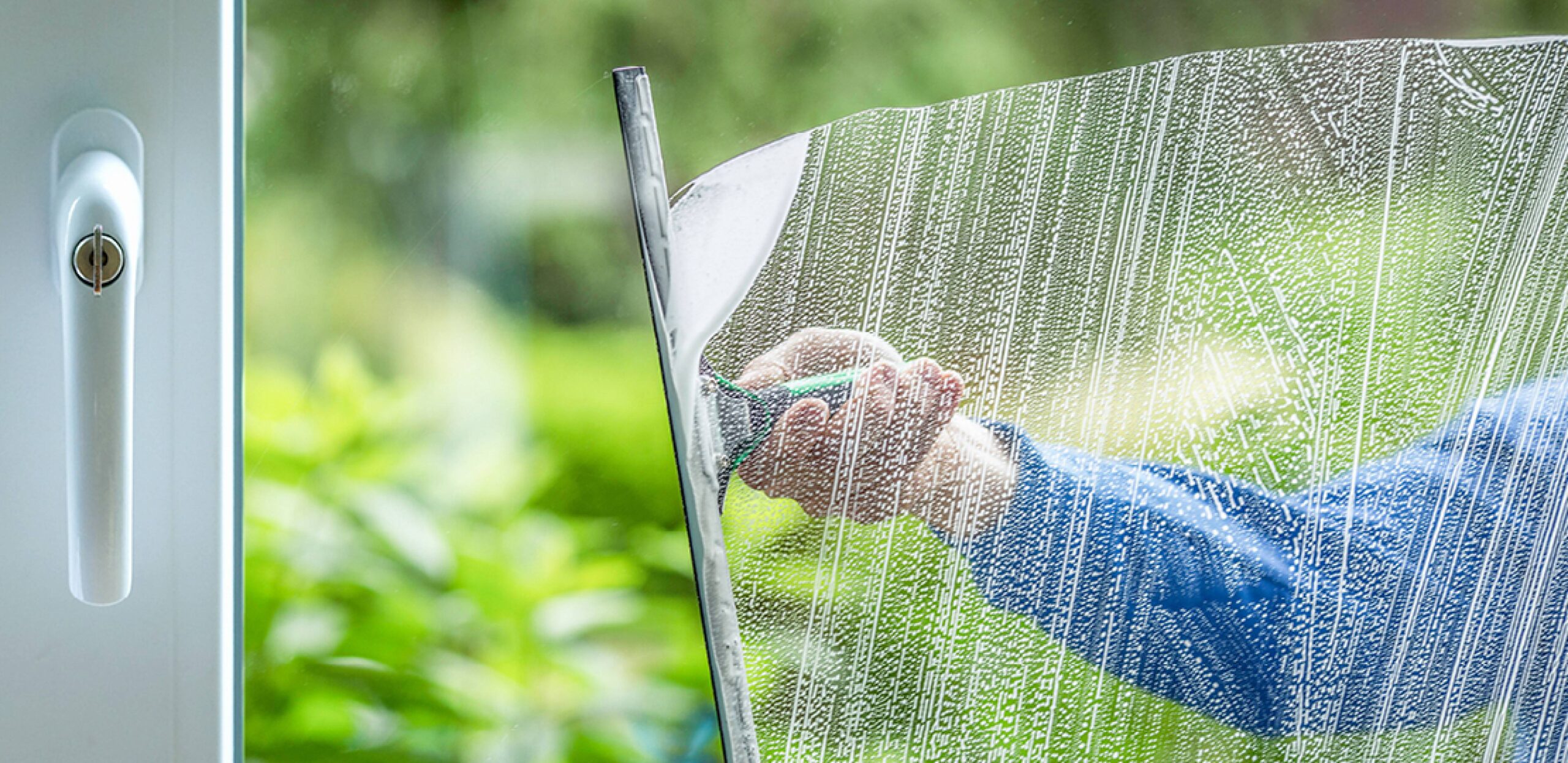 A professional window washer uses a squeegie to precisely clean a modern window, with a lush green yard in the background.