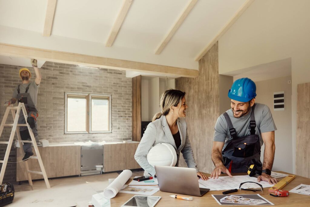 Female homeowner with Get Dwell contractor reviewing architectural plans in a kitchen undergoing a major remodeling with a carpenter on a ladder in the background