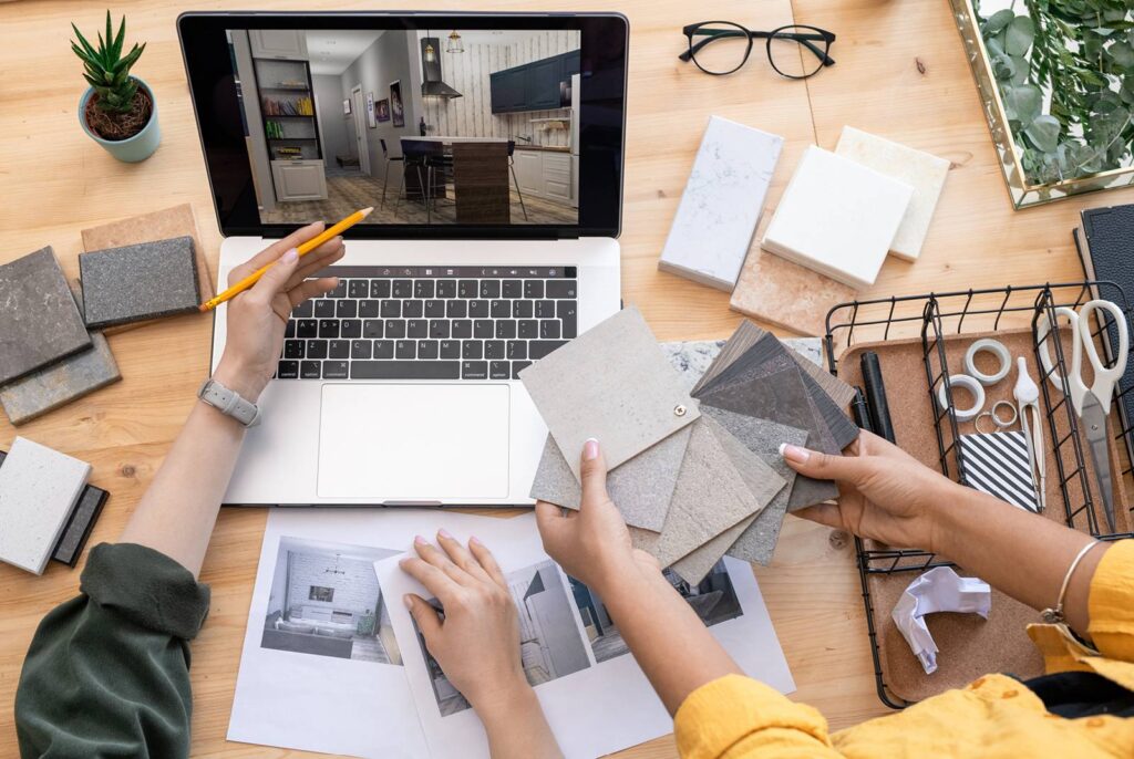 Two people are working on a remodeling project at a wooden table. One person is holding a pencil and pointing at a laptop screen displaying an interior design image, while the other person is holding various countertop samples. The table is scattered with different material samples, a small potted plant, eyeglasses, scissors, and other stationery items.