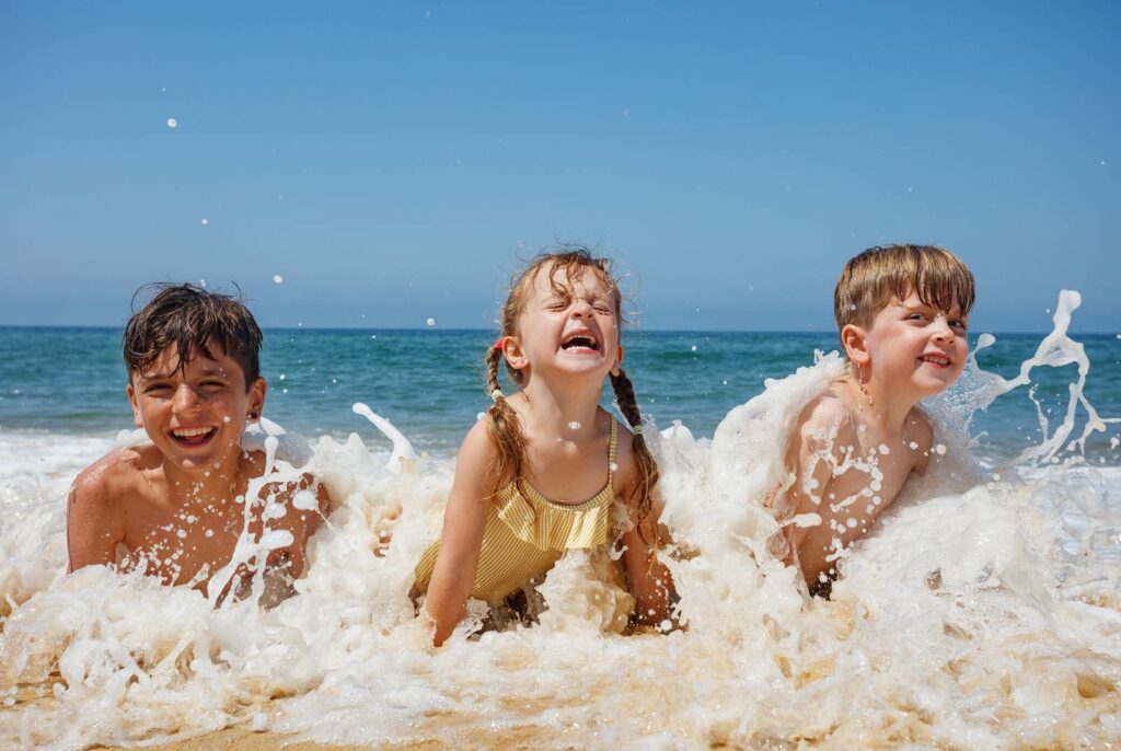 Three young children are gleefully laughing, splashing in water at the beach in the sand in the summer