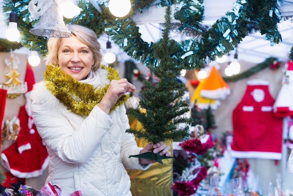 Woman in a white puffer jacket with a gold garland wrapped around her neck like a scarf holding a miniature artificial Christmas tree and an ornament with Holiday lights and decorations in the background of a local independent hardware.