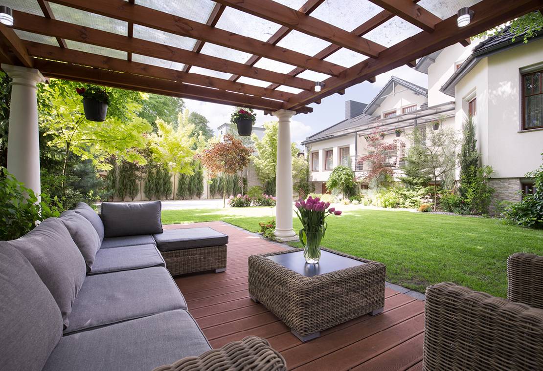 A beautiful wooden deck in the back yard of a stately stucco home, with comfortable cushioned couches and a beautiful table with a vase of flowers. The deck is shaded by custom pergola.