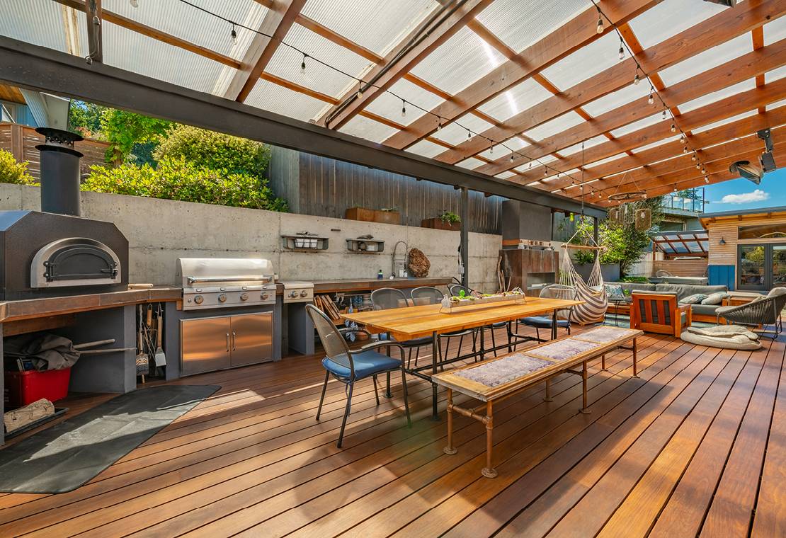 A large wooden deck with a wood burning oven, long wooden table with benches and chairs, a grill, a full outside kitchen, and a pergola overhead with translucent panels that are letting the sunlight in but protecting the deck.