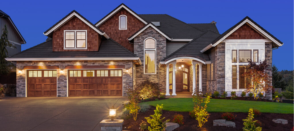 A large, impressive suburban home at dusk, with handsome light fixtures lighting the super clean driveway and illuminating the house and its perfectly manicured front lawn and garden.