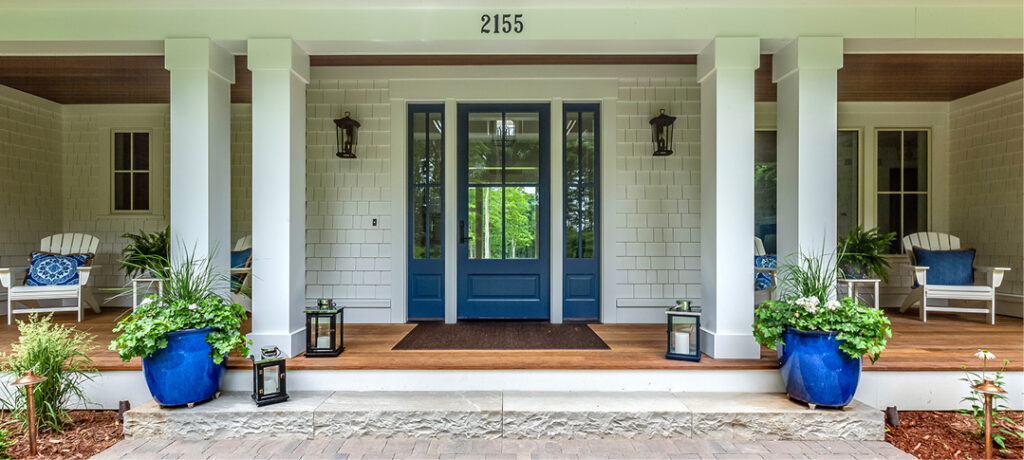 The front entry way to a white shingled home, with stately white columns, a pristine stone sidewalk and wodden deck, cobalt blue planters on either side of the entry way, crisp white adirondack chairs on either side of the porch with cobalt blue pillow, green wispy ferns and copper sidewalk lighting.