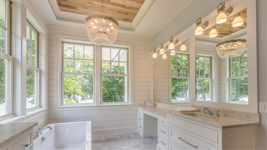A light filled bathroom with two walls of windows reflecting trees from a wall of cabinets with two large mirrors and charming glass and metal light fixtures. The walls, cabinet and ceiling are color drenched in a soft, warm grey and the walls are sided with shiplap. The ceiling has a recessed inset of warm wood paneling, from which hangs a graceful beaded chandelier. Below it is a freestanding bathtub, and grounding the space are light gray and white marbled tiles.