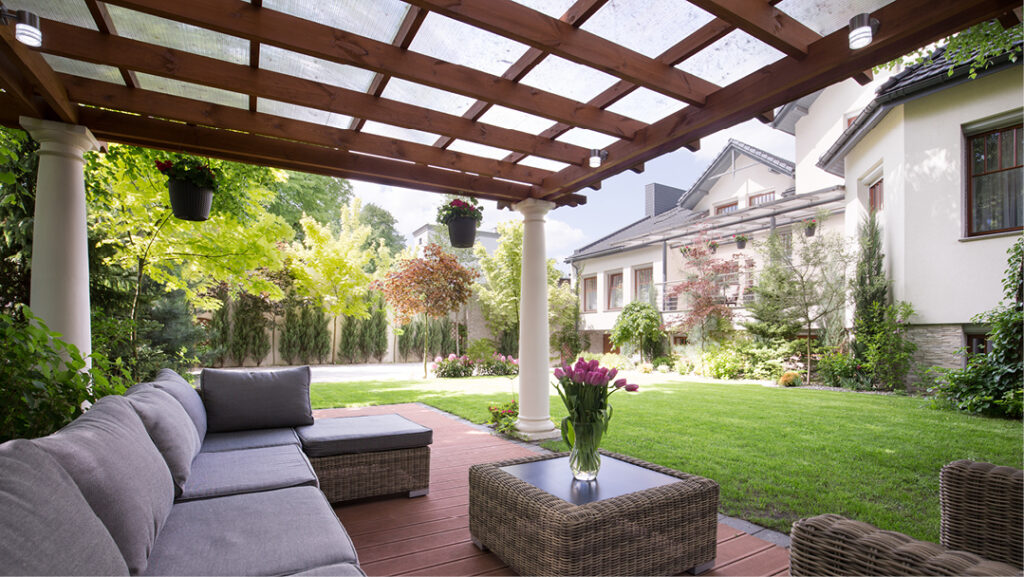A view from a wooden deck in a manicured back yard. A beautiful wooden pergola with translucent panels and large white columns overhangs a cushioned outdoor couch with wicker tables and chairs and a beautiful vase of pink tulips. We can see a large suburban home in the background, surrounded by trees in a variety of colors.