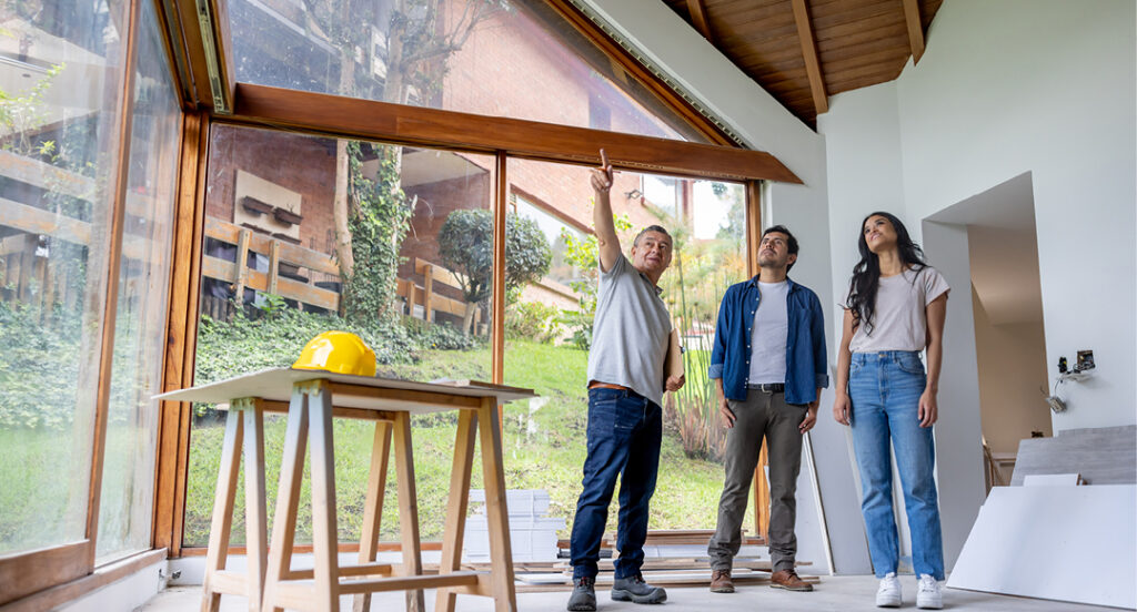 A young couple look dreamily up to where a smiling contractor with a clip board is pointing. Nearby, two saw horses hold a board and we can see a yellow hard hat. The room is walled with windows and has a wooden ceiling that angles up. Drywall panels are leaned against a wall.