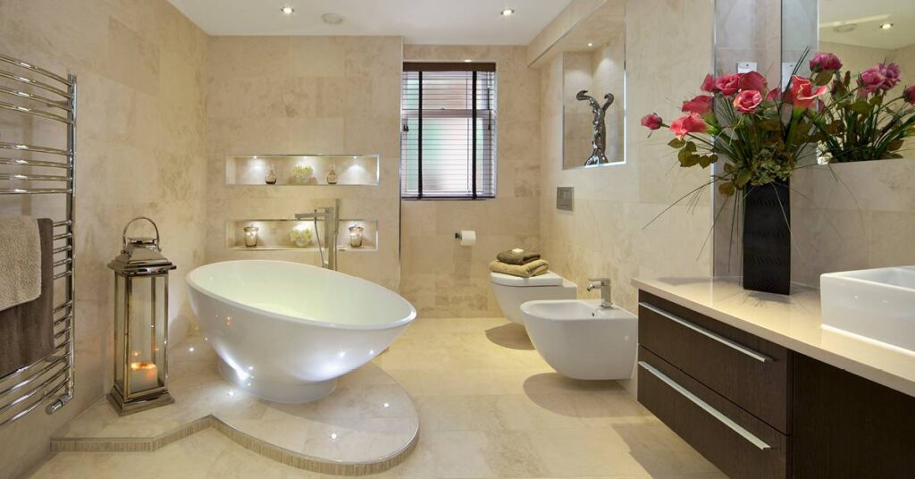 A warm, inviting bathroom with walls and floor enveloped in sandy marble tiles, a dark brown streamlined vanity with a black vase of dramatic burgundy roses, and a crisp, white, modern freestanding tub, toilet and bidet. The bathroom has been thoughtfully designed with inset tiled shelves and lighting that highlights are pieces. A silver towel warmer hangs just near the bathtub, and a large silver and glass lantern sits with the tub atop a custom sculptured raised area. The ceiling also has dimmable spot lighting, which altogether creates a warm and inviting space where you want to linger.