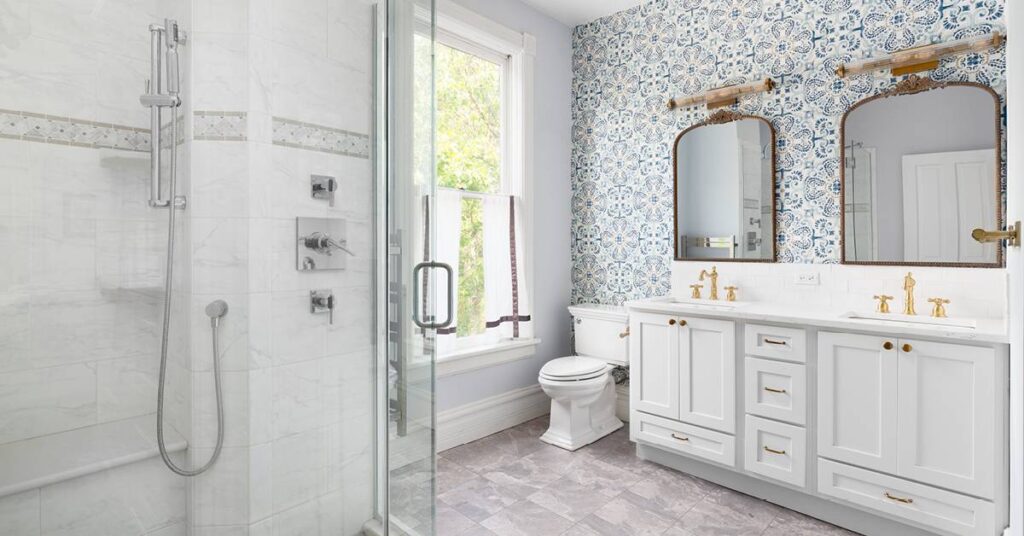 A bright, sun-lit bathroom with a double sink white vanity that boasts brass hardware and fixtures, two bronze mirrors with lighting, a blue and white tile wall, and gray and white shower tiles and flooring.