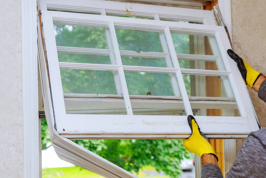 A Get Dwell installer wearing yellow gloves removes an old window frame from its wooden casing, with a lush green view outside. 