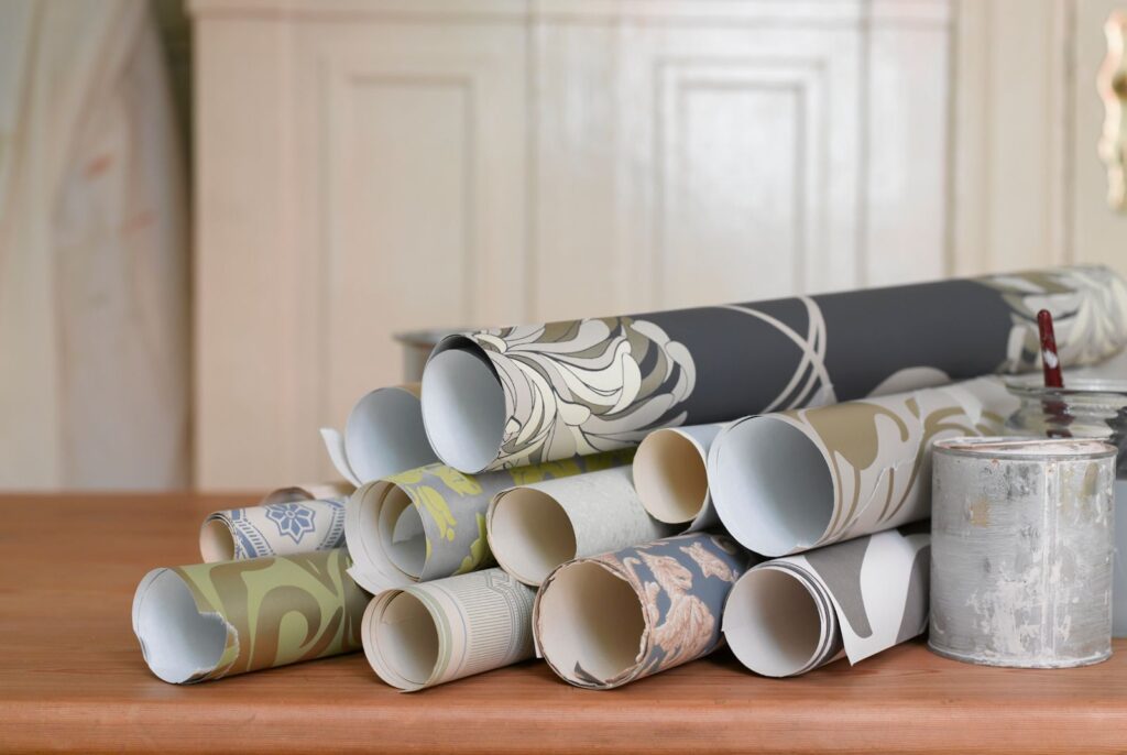 Rolls of high end, designer wallpaper are next to a wallpaper paste bucket on a rich wood table
