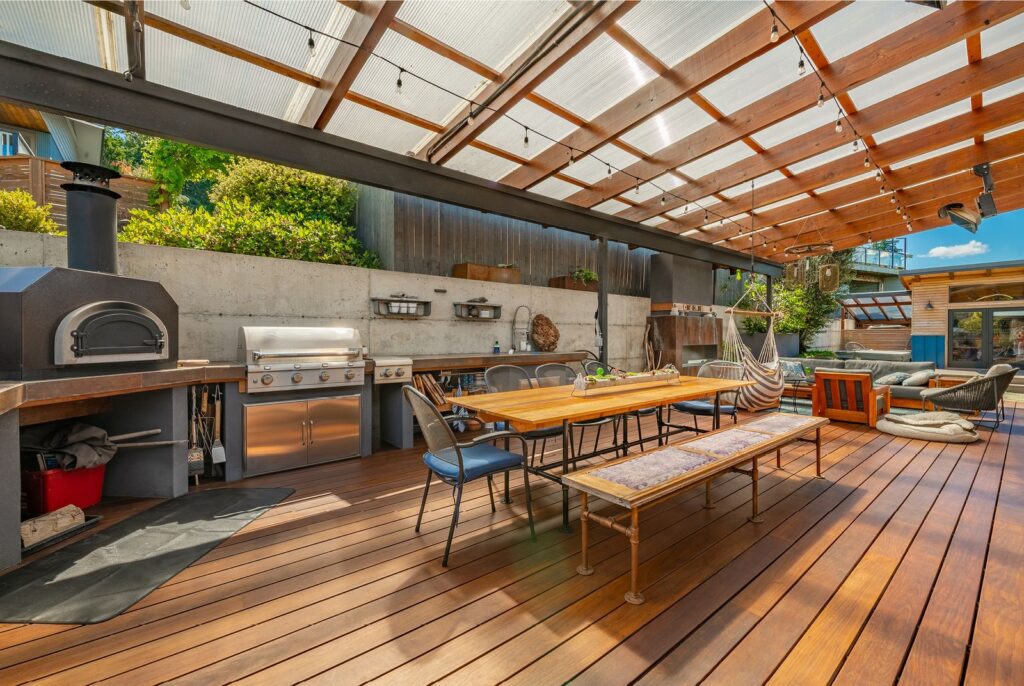 a large, covered wooden deck with a wood fired pizza oven, outdoor kitchen, huge table, and comfortable seating area with fireplace, ready for a Labor Day party