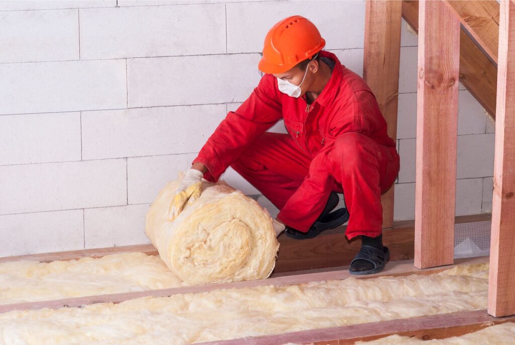Worker in red coveralls, hard hat and dust mask unrolls fiberglass insulation between attic floor joists.