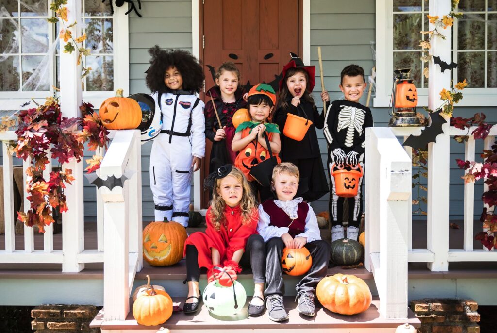 Kids in Halloween costumes hold candy buckets on a front porch decorated with jack-o'-lanterns and fall leaves.